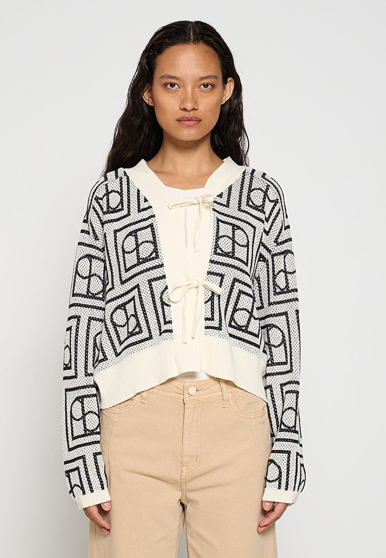 Schwarz-weiße, kurz geschnittene Strickjacke mit geometrischem Muster, Kordelzug am Halsausschnitt und langen Ärmeln. Getragen über einem hellen Oberteil.