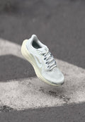 Hellgrauer Sportschuh mit Mesh-Obermaterial, reflektierenden Akzenten und gepolsterter Sohle, schwebend über einer grauen Oberfläche mit weißen Markierungen.