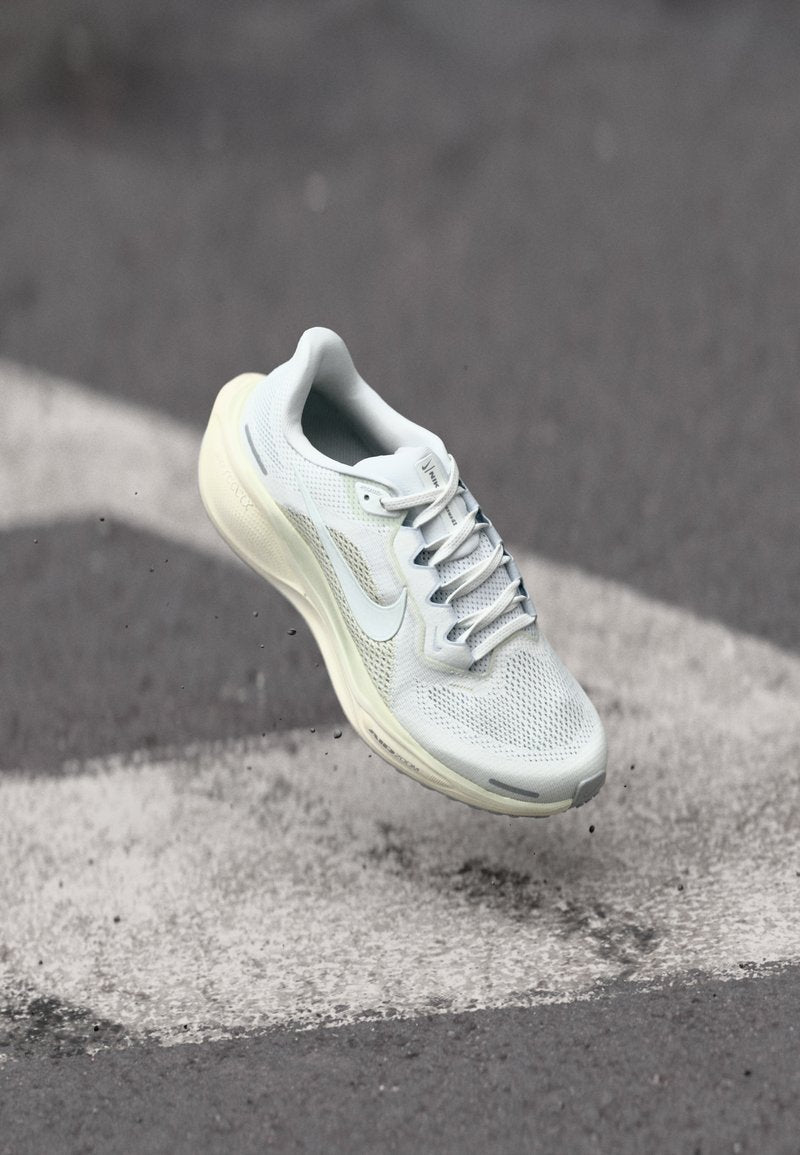 Hellgrauer Sportschuh mit Mesh-Obermaterial, reflektierenden Akzenten und gepolsterter Sohle, schwebend über einer grauen Oberfläche mit weißen Markierungen.