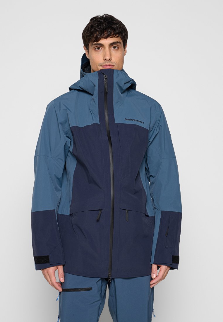 Mann trägt eine blaue und marineblaue wasserdichte Kapuzenjacke mit Reißverschluss und Klettverschlüssen an den Ärmelenden, steht vor einem einfarbigen Hintergrund.