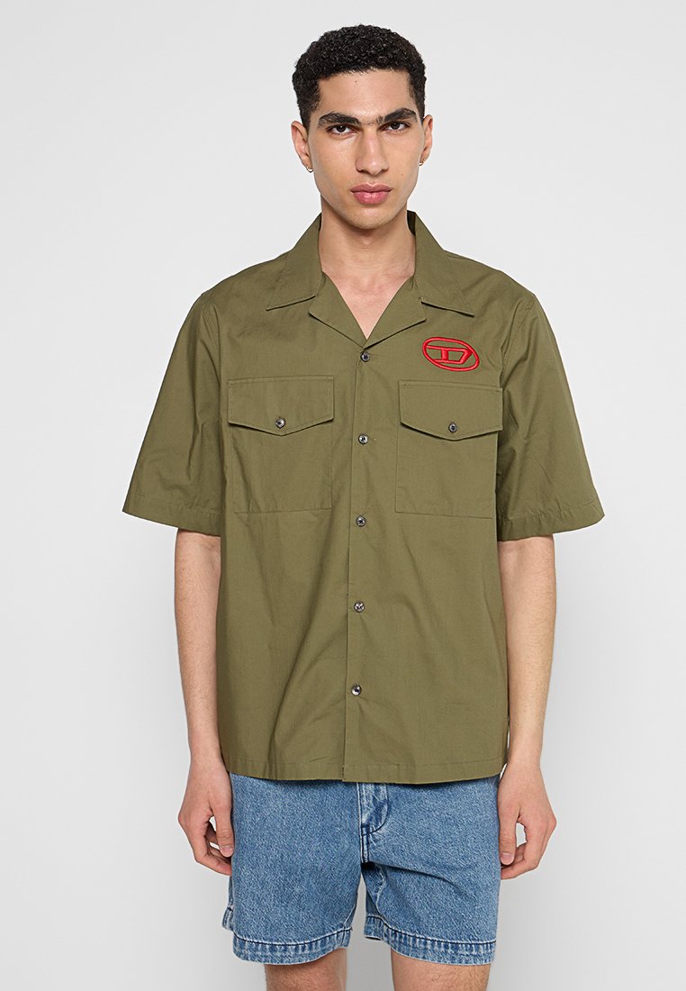 Olivenfarbene Kurzarmbluse mit Patch, zwei Fronttaschen, Knopfverschluss und strukturiertem Finish; kombiniert mit Jeans-Shorts.