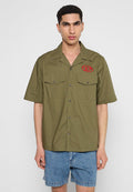 Olivenfarbene Kurzarmbluse mit Patch, zwei Fronttaschen, Knopfverschluss und strukturiertem Finish; kombiniert mit Jeans-Shorts.