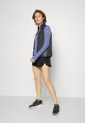 Junge Frau in schwarzem Sportwesten, blauer Langarmshirt, schwarzen Laufshorts und schwarzen Turnschuhen, die gegen eine weiße Wand steht.