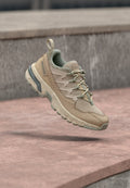 Beige und tanfarbene Salomon Trailrunning-Schuhe mit Obermaterial aus Mesh und Leder, punktiertem Sohlenmuster und grünen Akzenten, die über einer steinernen Oberfläche schweben.