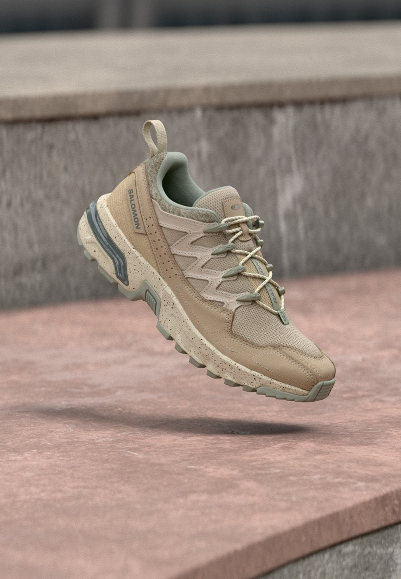 Beige und tanfarbene Salomon Trailrunning-Schuhe mit Obermaterial aus Mesh und Leder, punktiertem Sohlenmuster und grünen Akzenten, die über einer steinernen Oberfläche schweben.