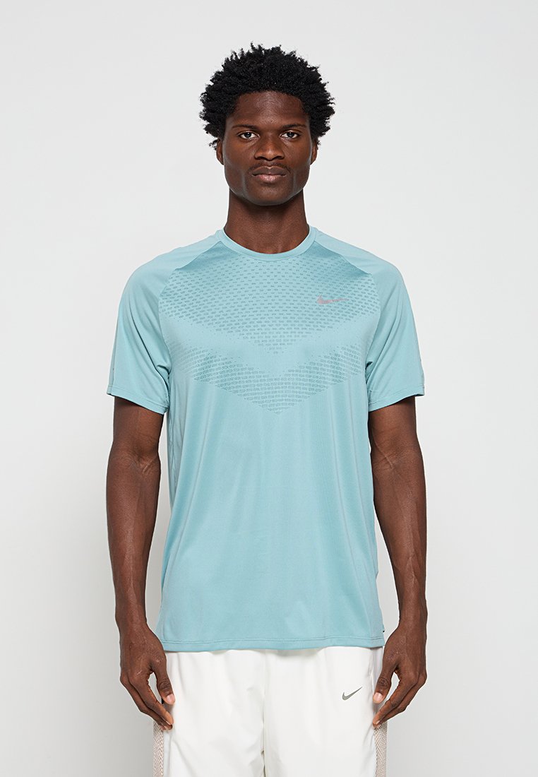 Mann mit kurzen lockigen Haaren, der ein hellblaues Sportshirt und weiße Shorts trägt, steht vor einem schlichten weißen Hintergrund.