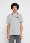Junger Mann mit lockigem Haar, der ein graues Poloshirt mit roten, weißen und blauen Streifen am Kragen und an den Ärmelenden trägt, steht vor weißem Hintergrund.