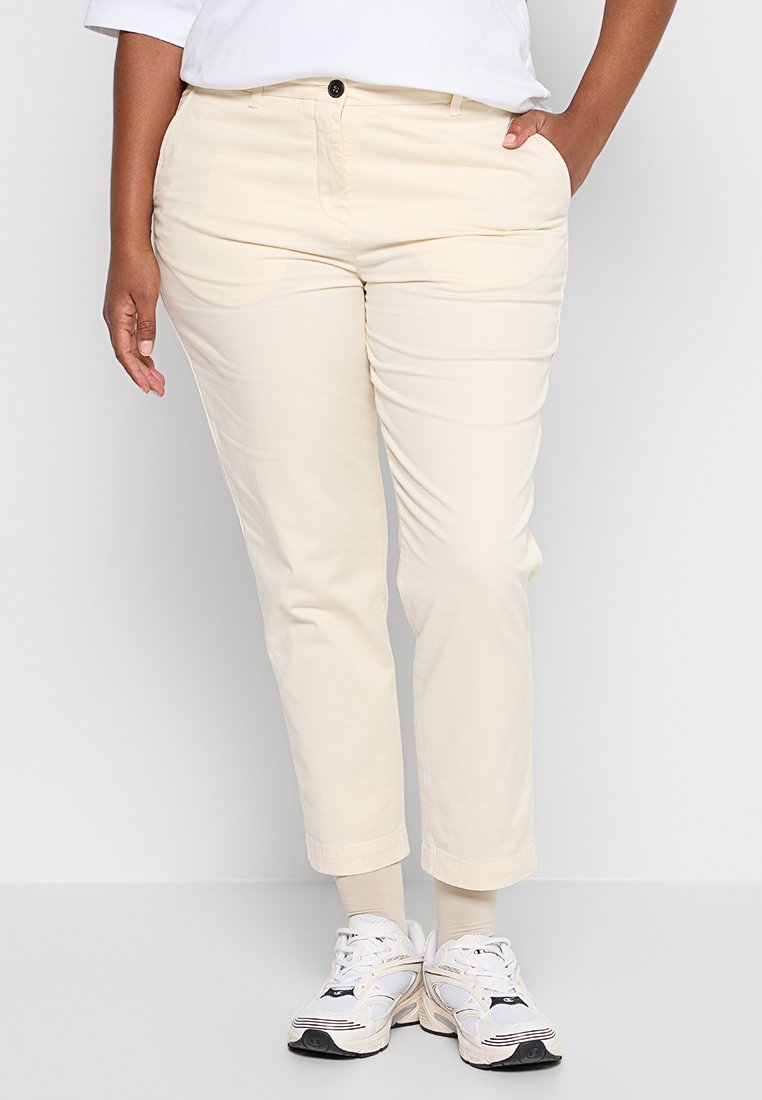 Beige Baumwollchinos mit schmaler Passform und Seitentaschen, abgeschlossen mit einem Knopfverschluss und umschlagenen Säumen, getragen mit weißen Sportschuhen.
