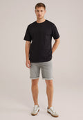 Junger Mann steht da und trägt ein schwarzes T-Shirt, graue hochgerollte Shorts und weiße Sneakers vor einem beigen Hintergrund.