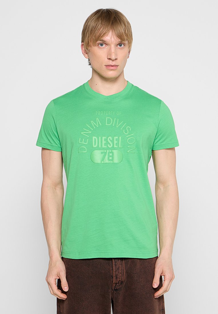 Grünes Baumwoll-T-Shirt mit Rundhalsausschnitt, das ein dezentes geprägtes Logo und eine lässige Passform aufweist, kombiniert mit dunkelbraunen Hosen.