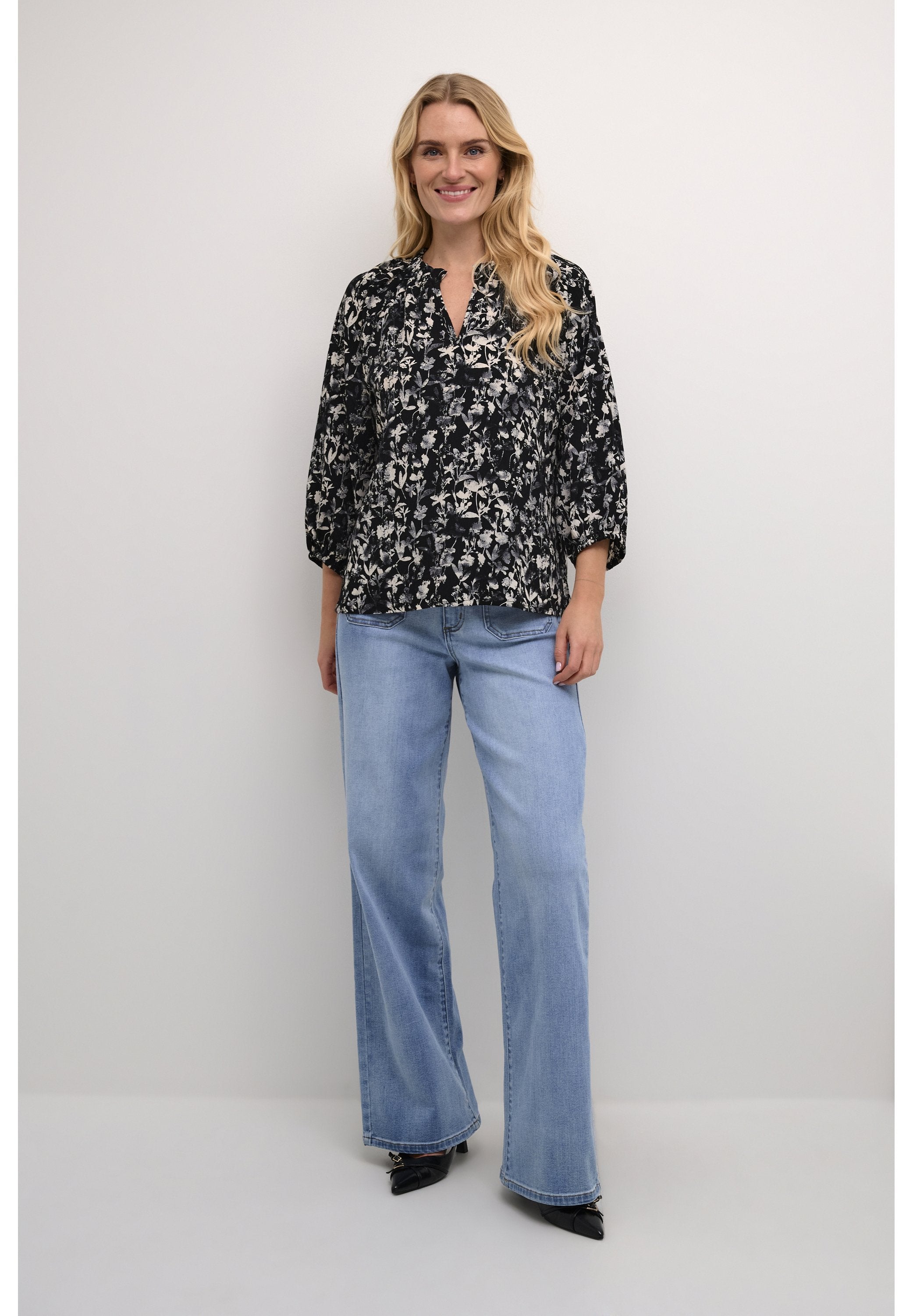 Schwarze Bluse mit floralen Mustern, lockerer Passform und Dreiviertelärmeln, kombiniert mit hellblauen weitem Beinkleid und spitzen schwarzen Schuhen.