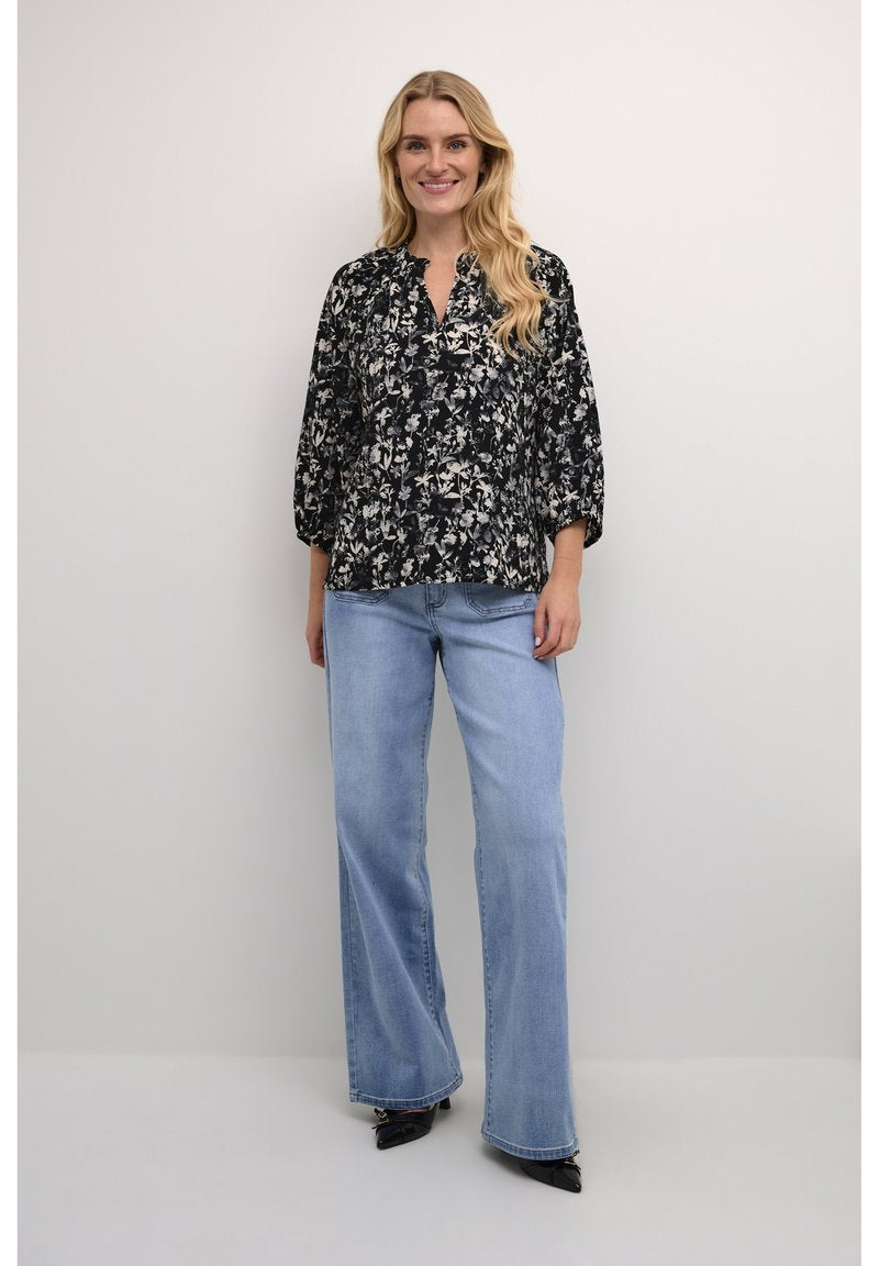 Schwarze Bluse mit floralen Mustern, lockerer Passform und Dreiviertelärmeln, kombiniert mit hellblauen weitem Beinkleid und spitzen schwarzen Schuhen.