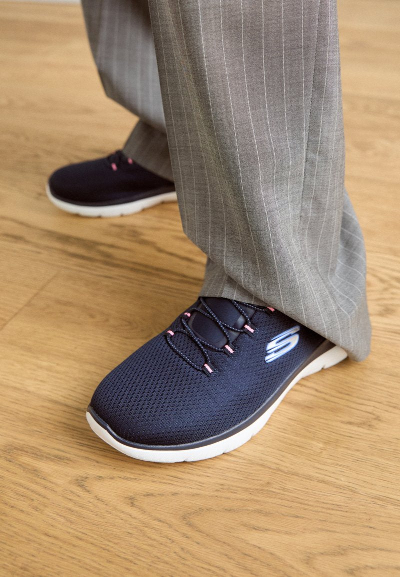 Marinefarbene Mesh-Sportschuhe mit rosa Akzenten und schwarzen Schnürsenkeln, ausgestattet mit einer weißen Sohle, kombiniert mit grauen schmal gestreiften Hosen auf einem Holzfußboden.