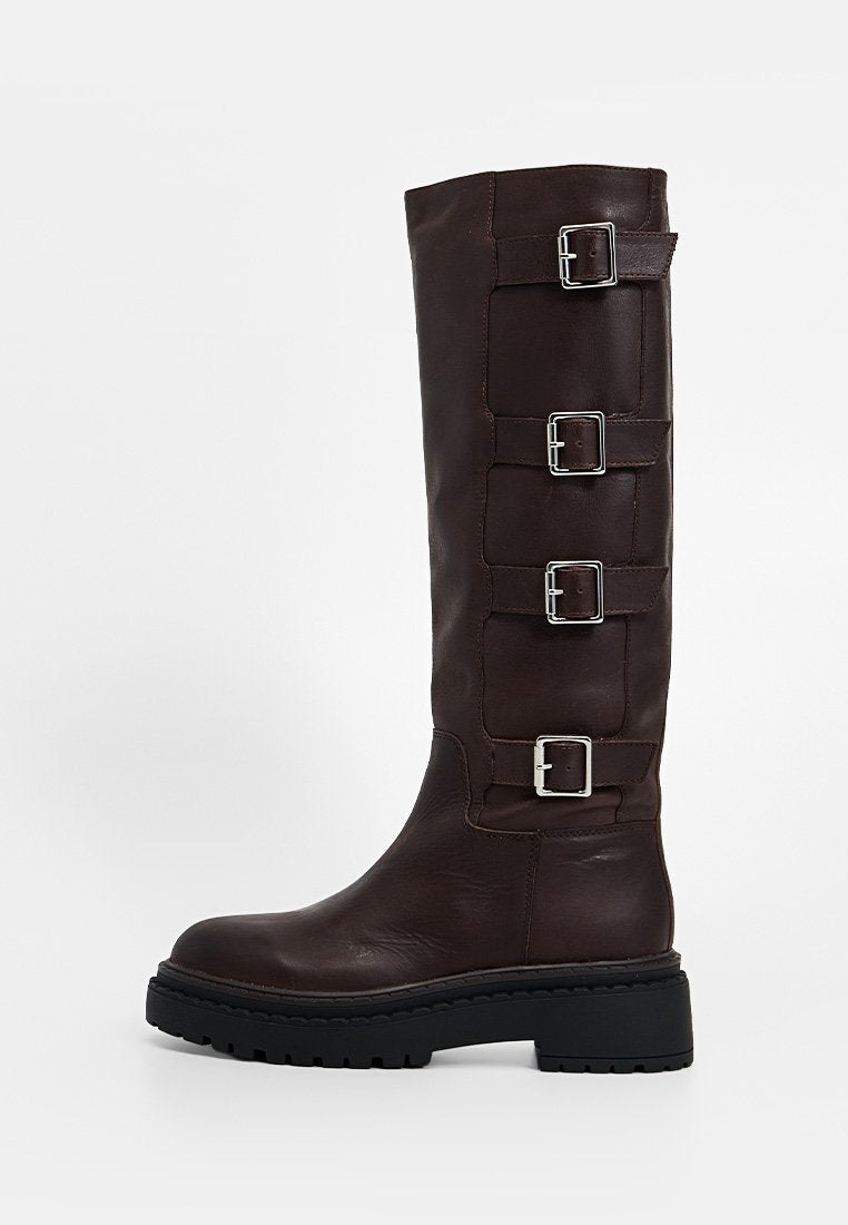 Hoher dunkelbrauner Lederstiefel mit vier silbernen Schnallen an der Außenseite und dicker schwarzer Profilsohle.