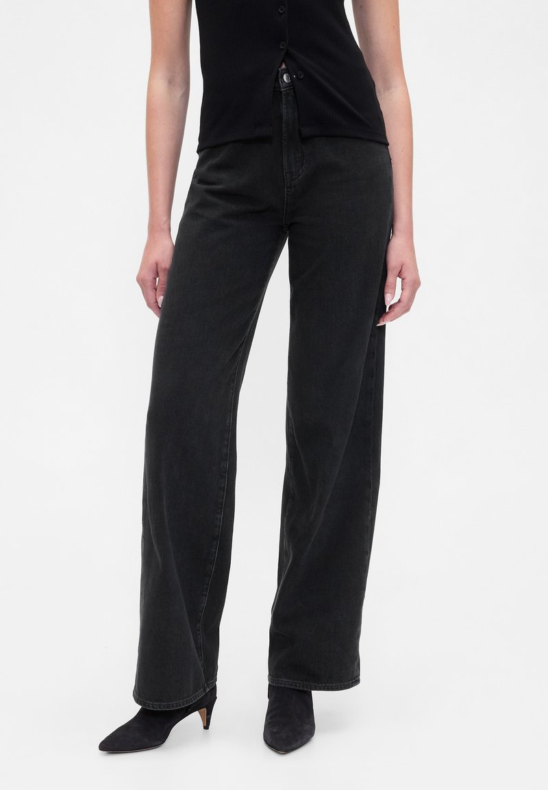 Schwarze Jeans mit weitem Bein aus Denim, hoch tailliert, mit Knopfverschluss und lockerem Schnitt, kombiniert mit spitzen schwarzen High Heels.