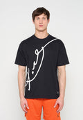 Mann trägt ein schwarzes, übergroßes T-Shirt mit großem weißem Schriftzug und orangene Basketball-Shorts, steht vor einem einfarbigen Hintergrund.
