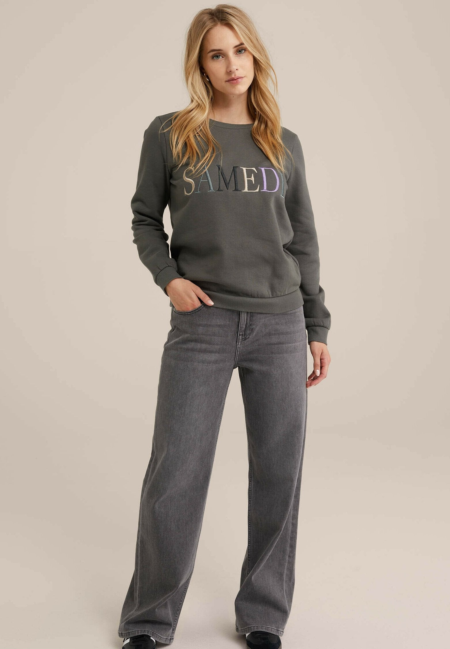 Grauer Sweatshirt mit dem Text "SAMEDI" in mehrfarbigem Stick, kombiniert mit hoch taillierten, weit geschnittenen grauen Jeans. Lässiger, bequemer Schnitt.