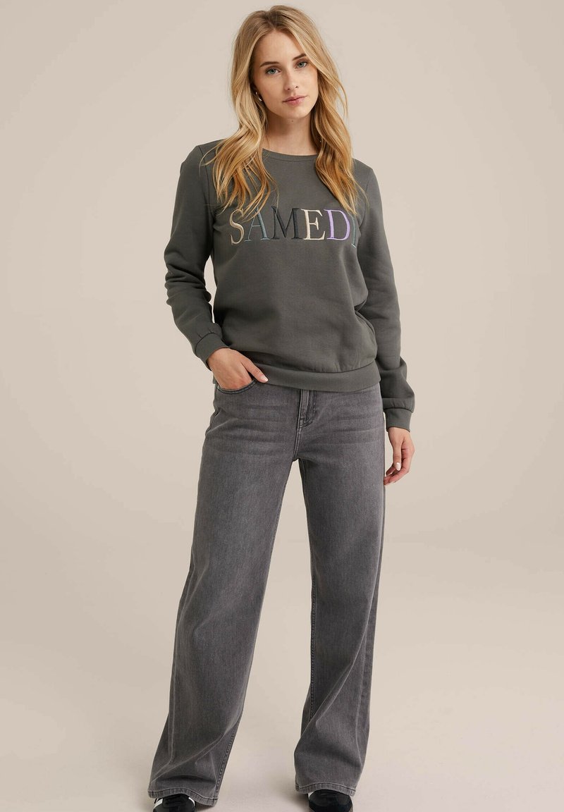 Grauer Sweatshirt mit dem Text "SAMEDI" in mehrfarbigem Stick, kombiniert mit hoch taillierten, weit geschnittenen grauen Jeans. Lässiger, bequemer Schnitt.