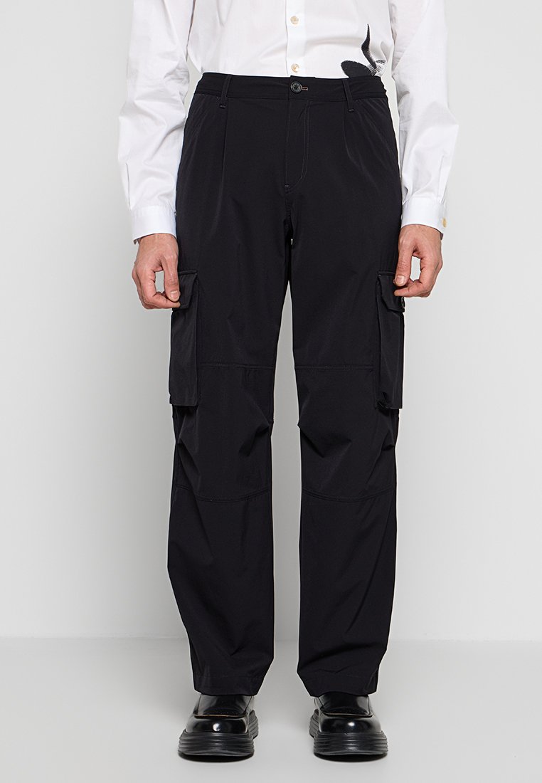 Mann trägt schwarze Cargo-Hose mit Seitentaschen, schwarze Schuhe und ein weißes Hemd mit Knöpfen und einem schwarzen Design auf der rechten Seite.