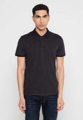 Junger Mann, der steht und ein schwarzes Poloshirt mit kurzen Ärmeln sowie dunkle blaue Jeans trägt, vor einem einfarbigen hellen Hintergrund.