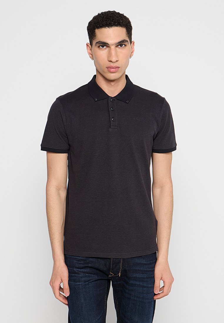 Junger Mann, der steht und ein schwarzes Poloshirt mit kurzen Ärmeln sowie dunkle blaue Jeans trägt, vor einem einfarbigen hellen Hintergrund.