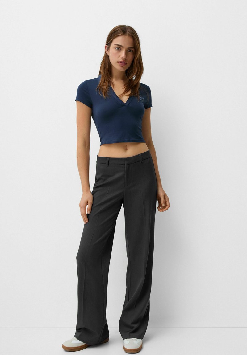 Marineblaues kurzärmliges Crop-Top mit V-Ausschnitt, kombiniert mit hochtaillierten, dunkelgrauen, weiten Hosen und hellgrünen Sneakern.