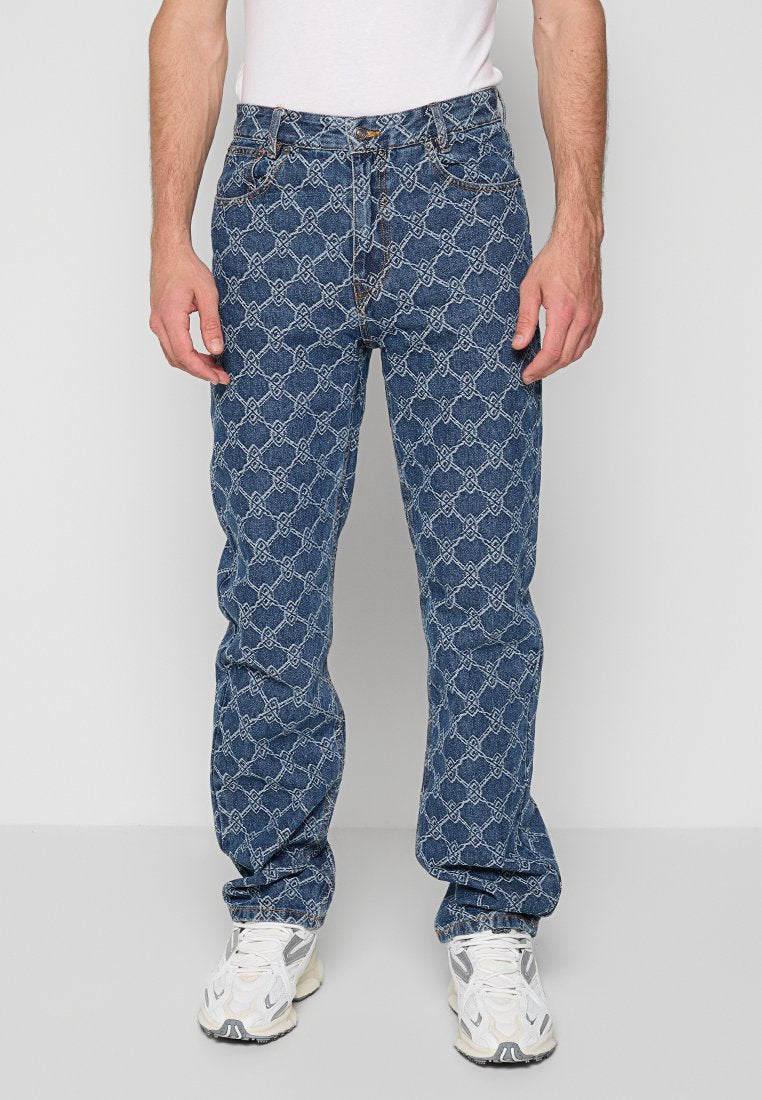 Mann trägt blue Jeans mit einem hellen geometrischen Kettenmuster, weiße Sneaker und ein weißes T-Shirt, steht vor einem schlichten Hintergrund.