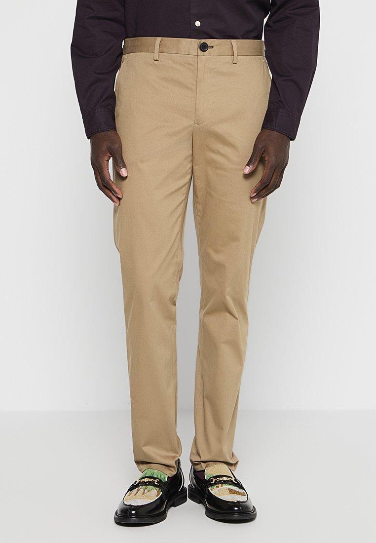 Beige Chinos mit geradem Schnitt und Gürtelschlaufen, mit Knopfverschluss und zwei Fronttaschen, kombiniert mit schwarzen verzierten Slippers.