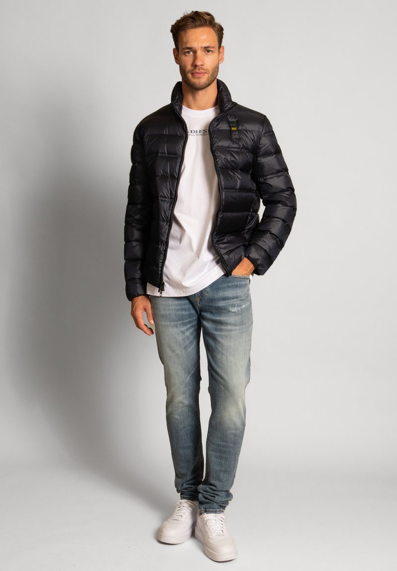 Schwarze gepolsterte Jacke mit hohem Kragen, Reißverschluss und gesteppter Textur, kombiniert mit einem weißen T-Shirt und hellblauen, ausgewaschenen Jeans.