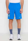 Mann trägt blaue Brasilien-Fußballshorts, weiße Sneakers, blaue Nike-Socken, steht vor einem einfarbigen hellen Hintergrund.
