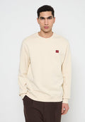 Cremfarbener Pullover mit gerippten Bündchen und Saum, der ein kleines rotes gesticktes Logo auf der Brust trägt. Einfaches, einfarbiges Design.