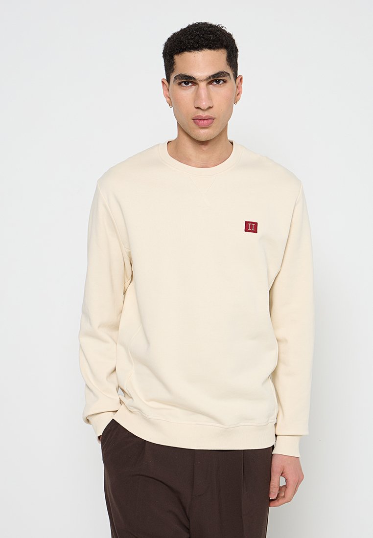Cremfarbener Pullover mit gerippten Bündchen und Saum, der ein kleines rotes gesticktes Logo auf der Brust trägt. Einfaches, einfarbiges Design.
