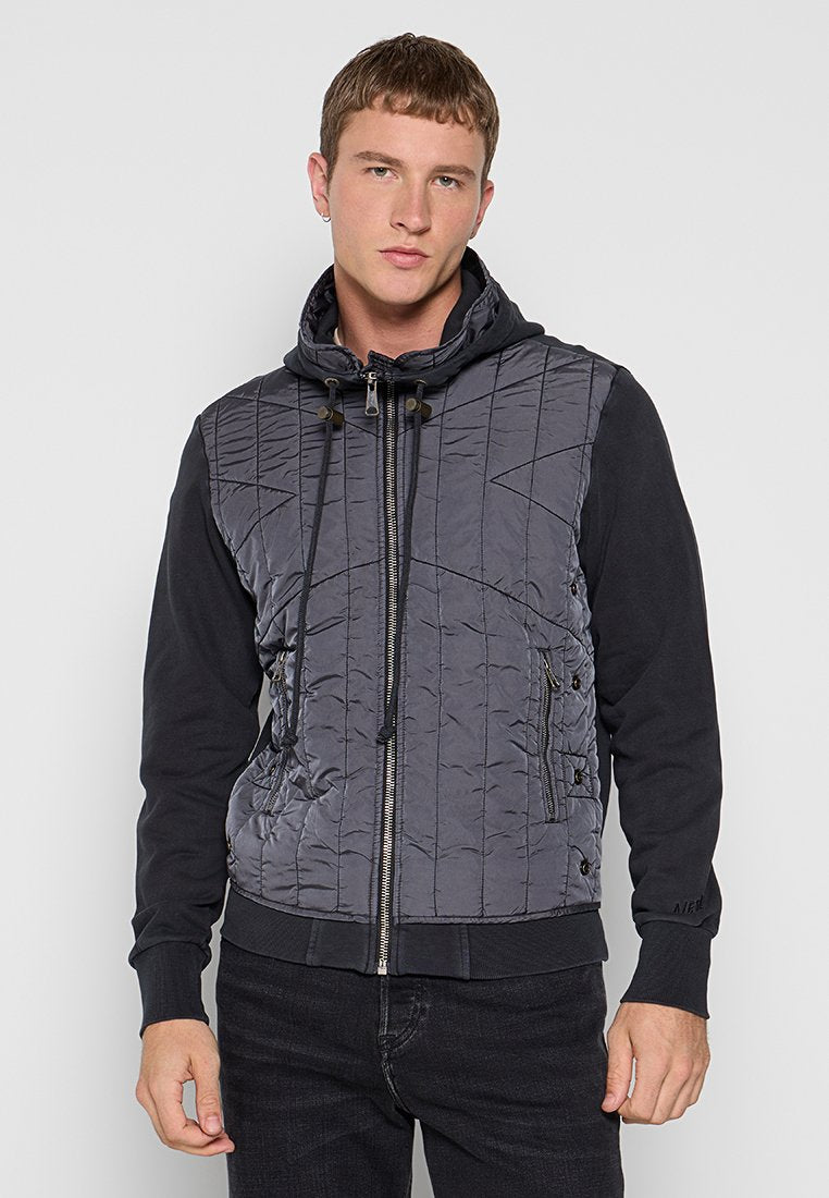 Junger Mann in einer schwarzen, gesteppten Kapuzenjacke mit Reißverschluss und Kordelzügen sowie schwarzen Ärmeln, der vor einem schlichten, hellen Hintergrund steht.
