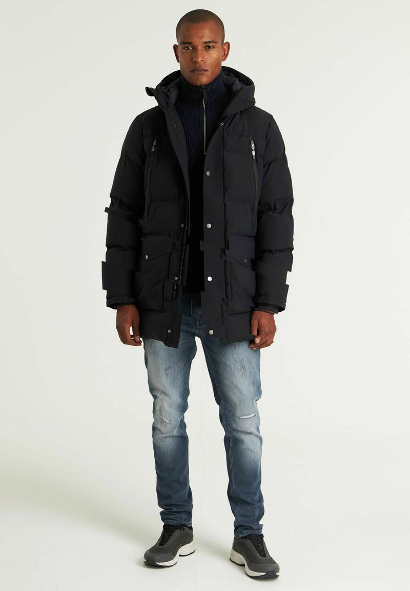 Schwarze gepolsterte Parka mit Kapuze, ausgestattet mit mehreren vorderen Taschen, Reißverschluss- und Druckknopfverschluss, getragen über einem dunklen Rollkragenpullover und hellblauen Jeans.