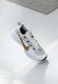 Weiße und graue Sneaker mit einem strukturierten goldenen Nike-Swoosh, weißen Schnürsenkeln und einer dicken weißen Sohle, die über einer reflektierenden Oberfläche schwebt.