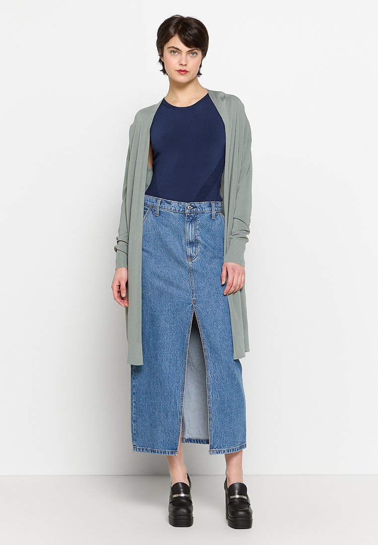 Langes Denimrock mit einem Schlitz vorne, kombiniert mit einem dunkelblauen Body und einer hellgrünen Strickjacke. Glänzende schwarze Plateauschuhe runden das Outfit ab.
