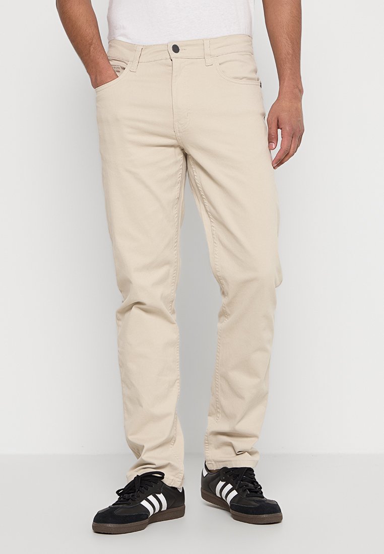 Beige Baumwollhosen mit geradem Schnitt, Fünf-Taschen-Design und Knopfverschluss. Paar schwarze Sneakers mit weißen Streifen.