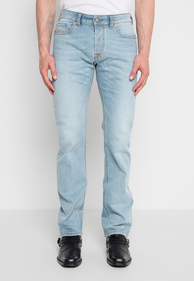 Mann trägt hellblaue Slim-Fit-Jeans und schwarze Lederschuhe, steht vor einem schlichten weißen Hintergrund.