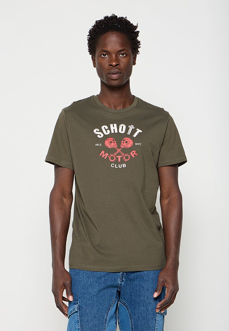 Junger Mann trägt ein olivgrünes T-Shirt mit "Schott Motor Club" und gekreuzten Totenkopf-Grafiken, kombiniert mit blauen Cargo-Jeans.