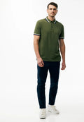 Ein Mann steht vor einem schlichten weißen Hintergrund, trägt ein olivgrünes Poloshirt mit marineblauen Abschlüssen, dunkelblaue Hosen und weiße Turnschuhe.