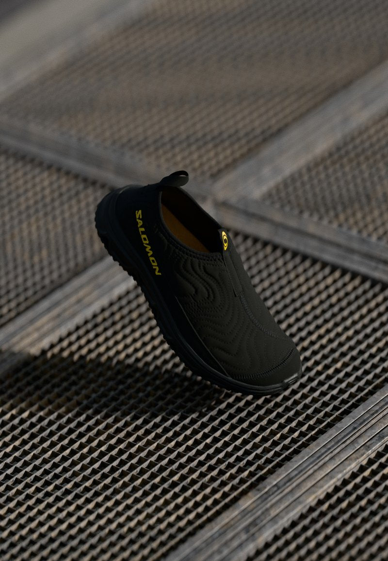 Schwarzer Slipper mit strukturierten Oberflächen, ausgestattet mit einem gelben Logo und einem Zugtab. Die Sohle ist für Traktion ausgelegt.