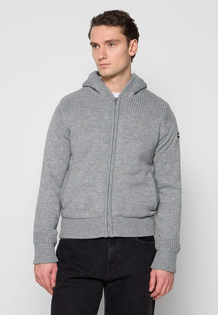 Grau gestrickter Kapuzenpullover mit durchgehendem Reißverschluss, gerippten Bündchen und Saum sowie einem strukturierten Muster an den Schultern. Darunter werden schwarze Hosen getragen.