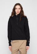Frau mit langem, welligem Haar, die einen schwarzen Pullover mit halbem Reißverschluss und beige Hosen trägt und vor einem einfarbigen hellgrauen Hintergrund steht.