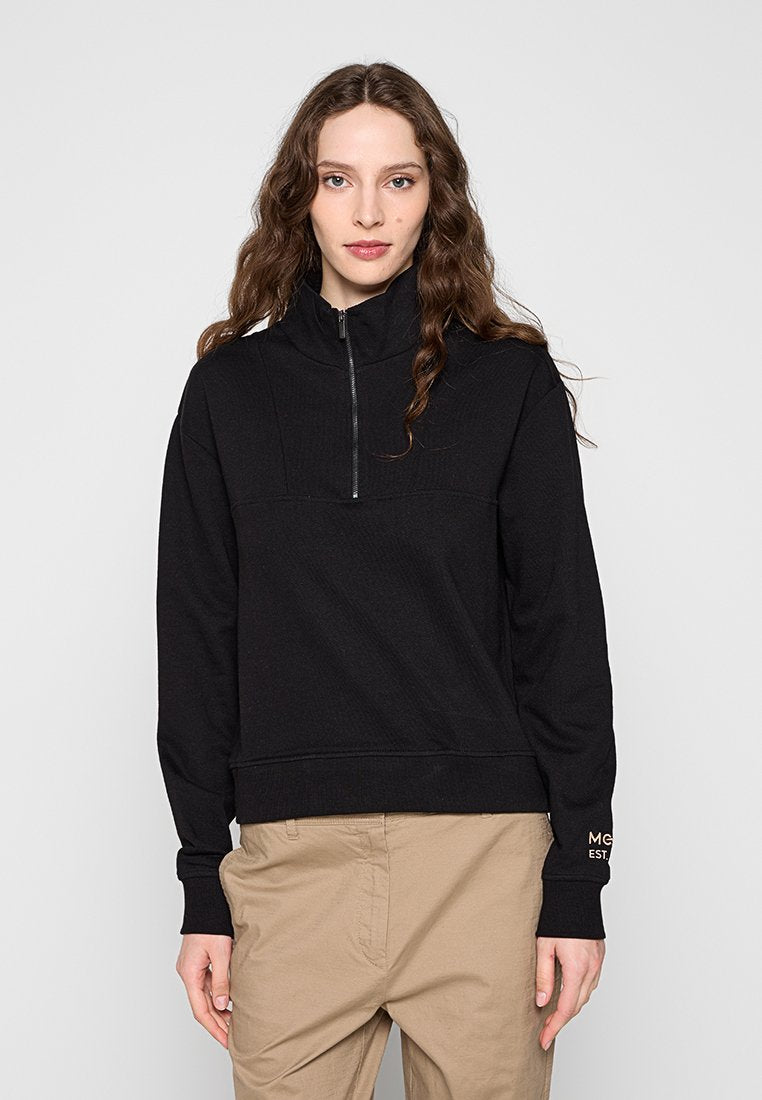 Frau mit langem, welligem Haar, die einen schwarzen Pullover mit halbem Reißverschluss und beige Hosen trägt und vor einem einfarbigen hellgrauen Hintergrund steht.