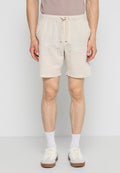 Mann trägt beige Shorts mit Kordelzug, weiße Socken und weiße Sneakers und steht vor einem einfarbigen hellgrauen Hintergrund.