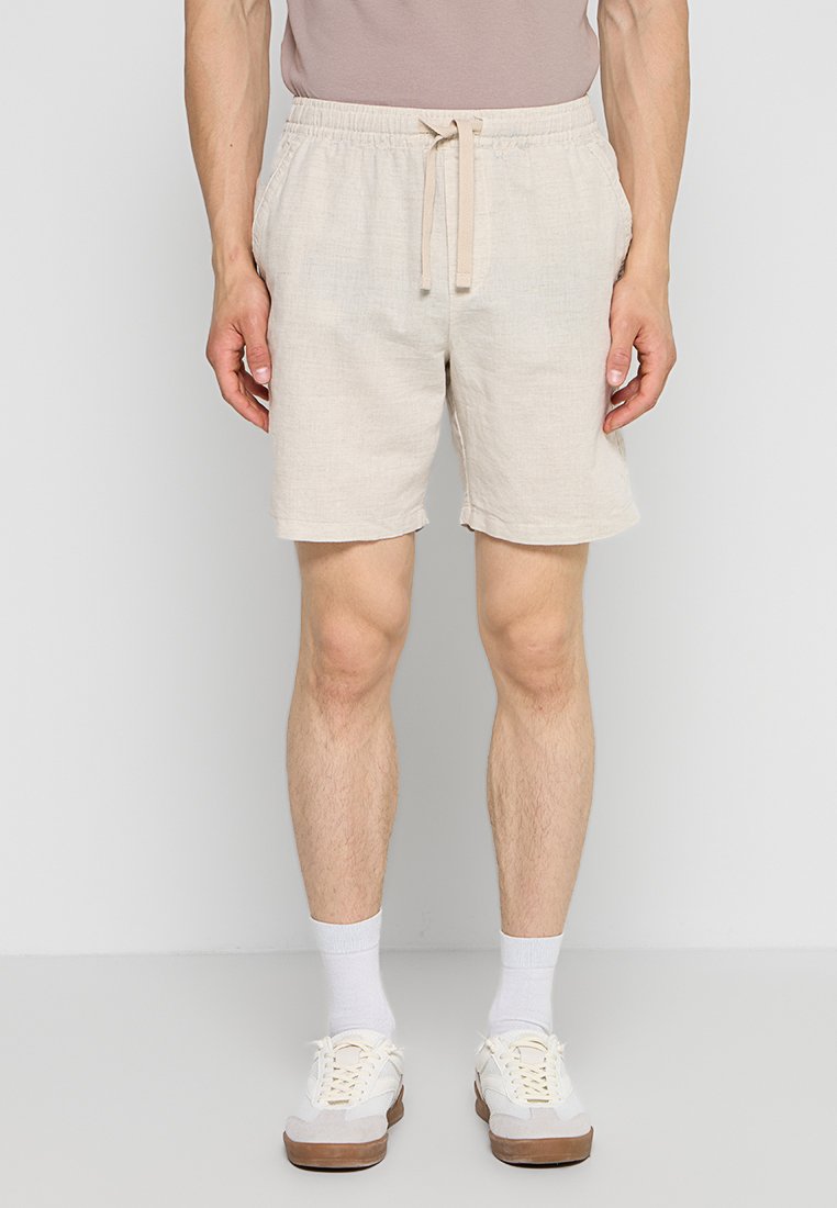 Mann trägt beige Shorts mit Kordelzug, weiße Socken und weiße Sneakers und steht vor einem einfarbigen hellgrauen Hintergrund.