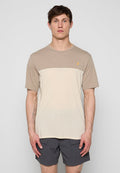Mann trägt beige und hellcremefarbenes T-Shirt mit Farbblockmuster und dunkelgraue Shorts, steht mit entspannt hängenden Armen vor einem schlichten hellen Hintergrund.