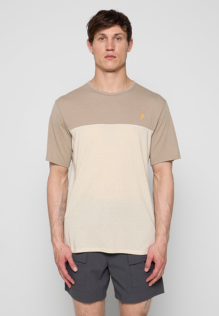 Mann trägt beige und hellcremefarbenes T-Shirt mit Farbblockmuster und dunkelgraue Shorts, steht mit entspannt hängenden Armen vor einem schlichten hellen Hintergrund.