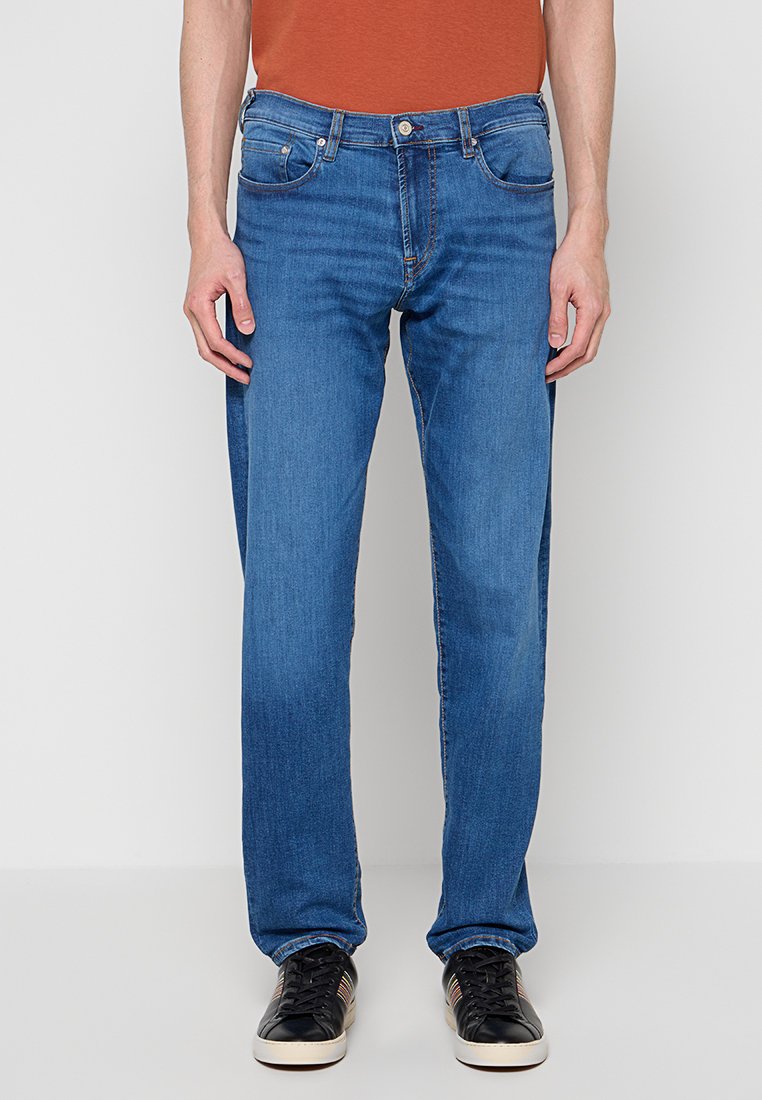 Blaue Jeans aus Denim mit geradem Bein, fünf Taschen und dezenter Naht. Kombiniert mit schwarzen Turnschuhen und einem rostfarbenen Hemd.