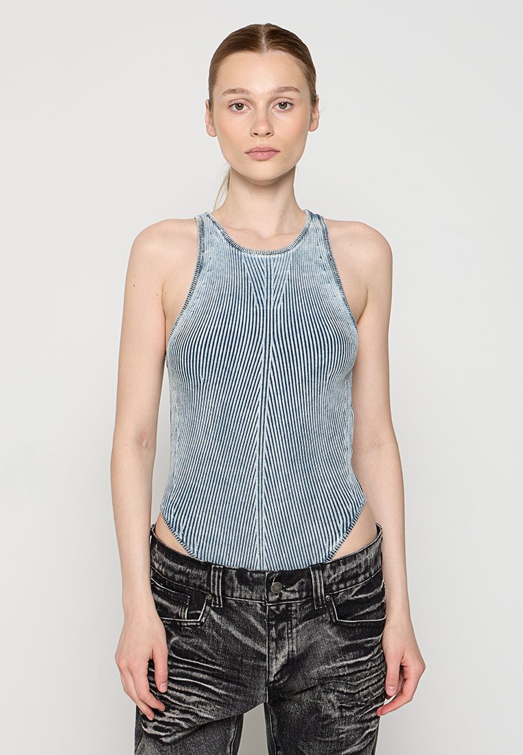 Frau mit hellen Haaren in einem ärmellosen, hochgeschlossenem, vertikal gestreiften blau-weißen Oberteil und schwarzen Acid-Wash-Jeans vor einem schlichten grauen Hintergrund.